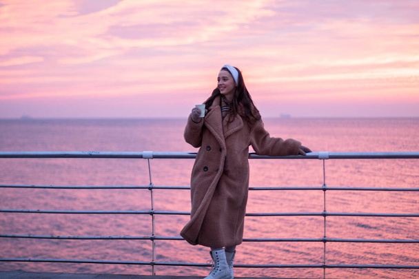 Una joven con un abrigo de piel con una taza caliente está cerca de la barandilla en el paseo marítimo, un amanecer rojo en el fondo
 - Foto, imagen