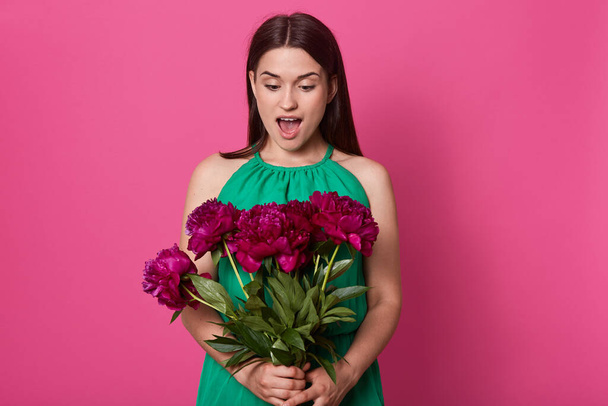 Beautiful cheerful girl stands isolated over pink studio background, holding bouquet of burgundy peony flowers, having astonished facial expression, poses with widely opened mouth. Surprise at 8 March - Фото, изображение