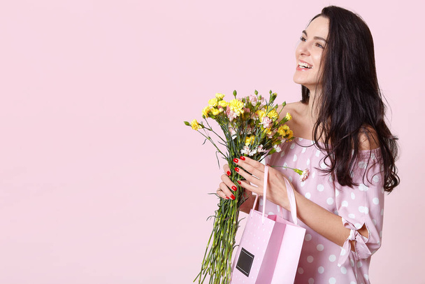 Horizontal studio shot of cheerful magnetic positive female looking aside, wearing dress, smiling sincerely, hoding flowers and bag with present, womens day concept. Copyspace for advertisement. - Foto, imagen