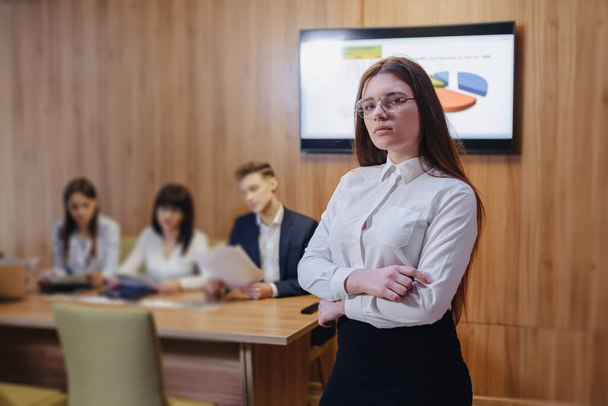 jovem atraente menina trabalhador de escritório elegante em óculos com um caderno nas mãos no fundo embaçado de colegas de trabalho
 - Foto, Imagem