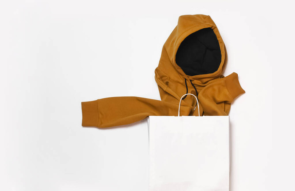 Yellow fashionable men's sweatshirt with a hood sticks out of white paper bag on white background top view. Fashionable male clothing, hoodie, casual youth style, sports. Hoody for design mockup. - Photo, Image