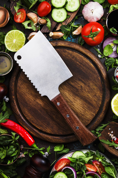 Healthy vegan ingredients layout. Flat-lay of green herbs, tomatoes, cucumbers, spices and oil on brown kitchen table with wooden board and hatchet knife. Top view, copy space. Vegetarian, raw, clean eating concept - Foto, imagen