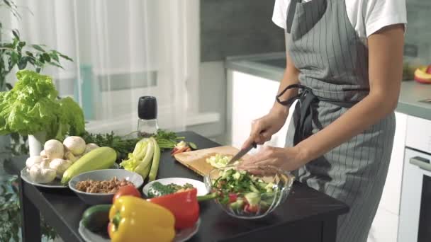 Hands of a young woman chopping fresh celery for salad on a wooden board and folding in a bowl to other vegetables in the kitchen. - Imágenes, Vídeo