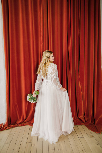 young beautiful blonde wedding bride in a beautiful white dress stands on a red background - Fotó, kép