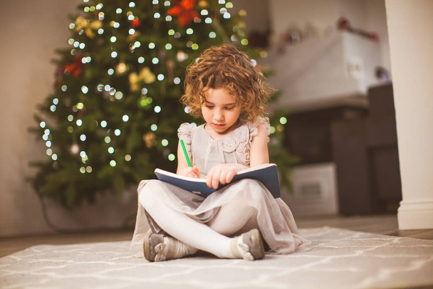 Preschool girl sitting next to festive tree and drawing - Foto, afbeelding