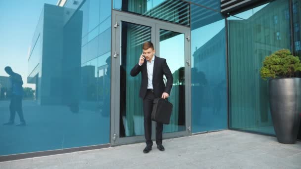 Young businessman with briefcase talking on phone and starts to run. Confident guy late for meeting. Office worker in suit runs near modern building. Handsome man in hurry to appointment. Slow motion - Footage, Video