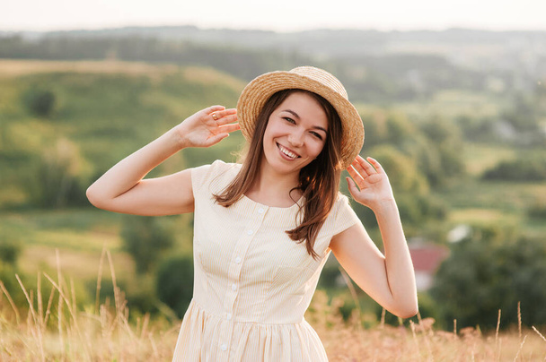 Brunette girl in a yellow dress and a hat on nature. Walks on a field on a hill. - Foto, Imagem
