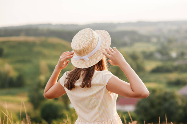 Brunette girl in a yellow dress and a hat on nature. Walks on a field on a hill. - Фото, зображення