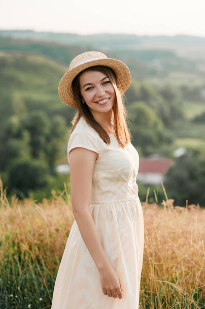 Brunette girl in a yellow dress and a hat on nature. Walks on a field on a hill. - Foto, imagen