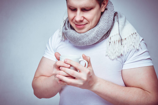 Мужчина в белой футболке с шарфом пьет вкусный чай от простуды и гриппа. Тонко. Закрыть
. - Фото, изображение