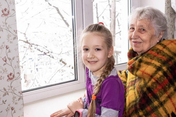 Happy beautiful great grandmother and her adorable granddaughter sitting at home with big window at winter - Photo, Image