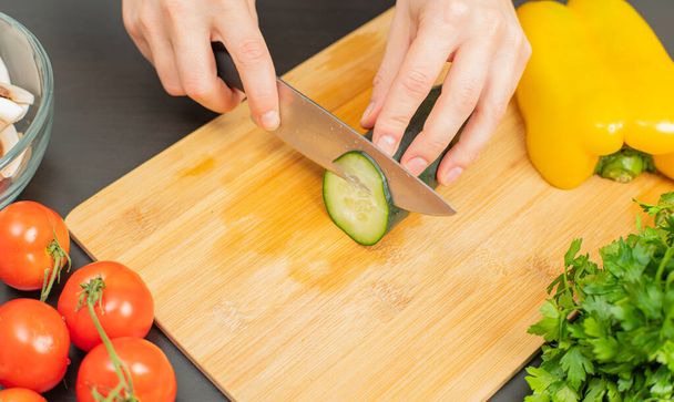 Woman is cutting cucumber on cut board on kitchen. Fresh vegetables for salad - Фото, изображение