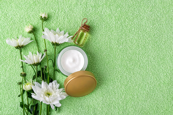 Face cream in an open jar with golden lid, bottle of chrysanthemum essential oil and white chrysanthemums on a green terry towel. Beauty, skincare and cosmetology. Copy space. Flat lay. - Foto, Imagen