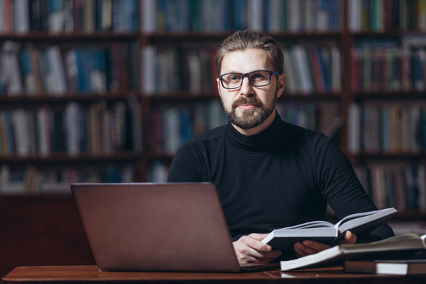 Człowiek z brodą siedzący w bibliotece z książkami i laptopem - Zdjęcie, obraz