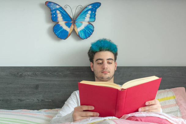 Mladý pohledný muž s modře obarvenými vlasy, čte si v posteli knihu v červeném, s vážným, soustředěným obličejem - Fotografie, Obrázek