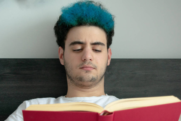 Detailní záběr dospívajícího s modrou barvou na vlasy, jak čte červenou knihu, velmi soustředěnou, zatímco leží na posteli - Fotografie, Obrázek