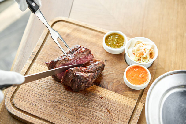The waiter is butchering griled meat with a knife. Grilled fillet steaks with Pickled cabbage and two sauces. Serving on a wooden Board. Barbecue restaurant menu. - Photo, Image