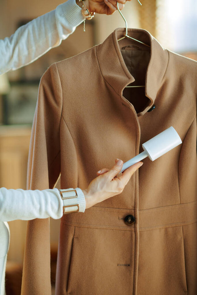 Preparing for cold season. Closeup on woman in white sweater cleaning coat on hanger with lint roller in the house in sunny winter day. - Foto, imagen