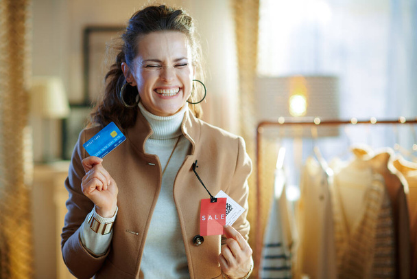 excited 40 years old woman in white sweater and skirt trying beige coat with red sale price tag and holding blue credit card in modern fashion store. - Photo, image