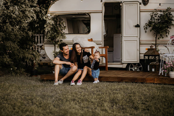 a young family meets the sunset near their mobile home, dad, mom and daughter, a young girl holds her daughter in her arms, her husband sits nearby and hugs his wife, the family travels - 写真・画像