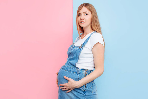 A beautiful young pregnant emotional woman in a denim jumpsuit poses on an isolated pink and blue background - 写真・画像