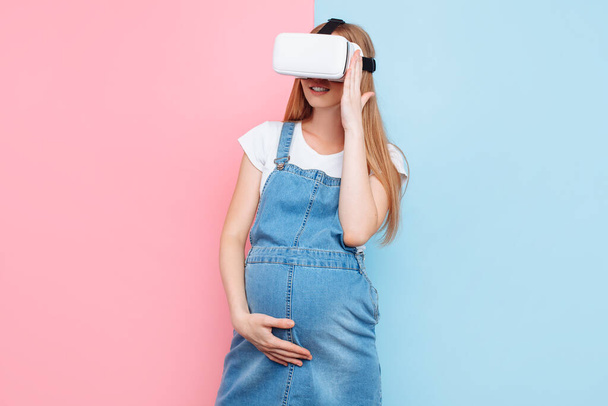 A cheerful happy pregnant woman has fun wearing virtual reality glasses while standing on a pink and blue background - Φωτογραφία, εικόνα