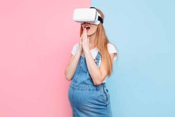 A cheerful happy pregnant woman has fun wearing virtual reality glasses while standing on a pink and blue background - Fotografie, Obrázek