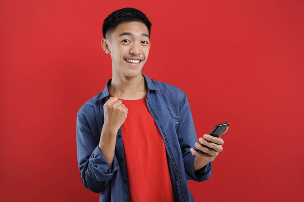 młody Azji nastolatek robi zwycięski gest trzymając telefon komórkowy, szczęśliwy dostać specjalny prezent online, izolowany na czerwonym tle - Zdjęcie, obraz