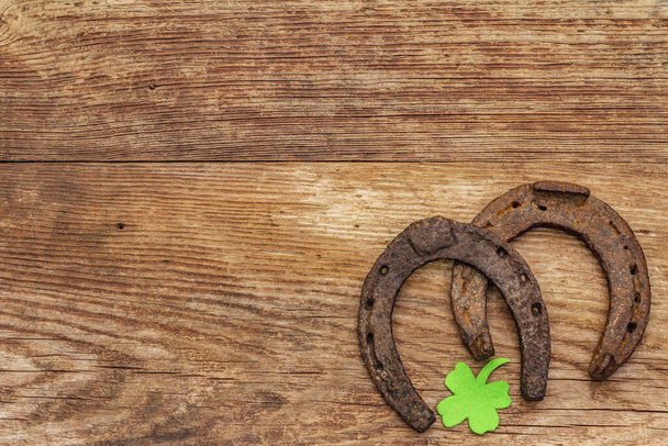 Two cast iron metal horse horseshoes, felt clove leaf. Good luck symbol, St.Patrick's Day concept. Old wooden background, horse accessories, top view - Photo, Image