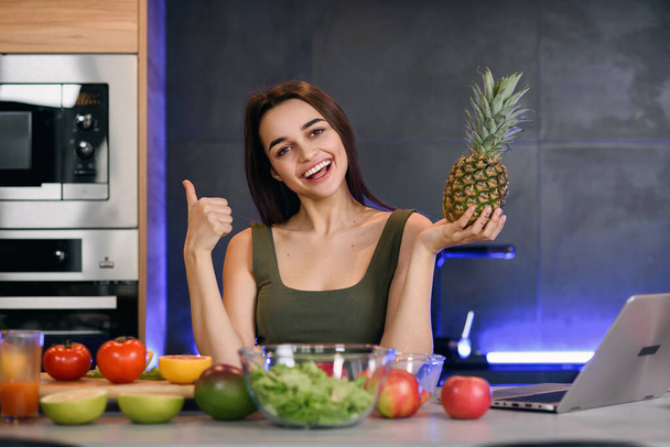 Healthy food at home. Happy woman is showing pineapple in modern kitchen. Vegan and vegeterian lifestile. - Φωτογραφία, εικόνα