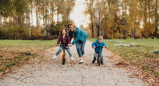 Urocza matka rasy kaukaskiej z dwójką dzieci, które z radością jeżdżą na rowerze w parku podczas spaceru - Zdjęcie, obraz