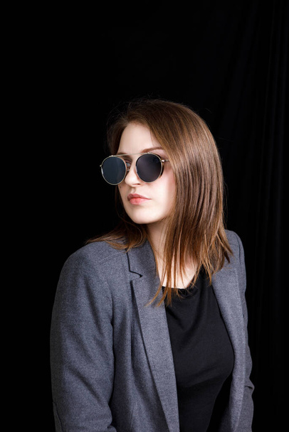 Imagen de una linda joven confiada de pie sobre fondo negro en el estudio, con chaqueta gris y gafas de moda, con el pelo rubio
 - Foto, imagen