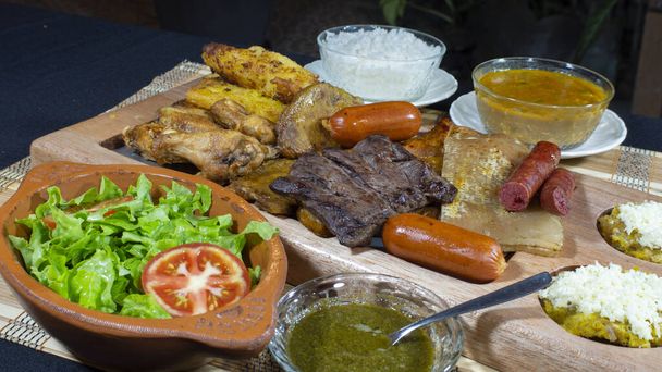 Μεγάλο ξύλινο δίσκο με ψητά κρέατα και λουκάνικα συνοδευόμενα από σάλτσες, τηγανητά κασάβα, ένα πιάτο ρύζι και σαλάτα ντομάτας και μαρουλιού σε μαύρο τραπέζι - Φωτογραφία, εικόνα