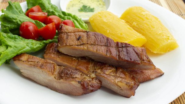 Λευκό πιάτο με καπνιστό χοιρινό κρέας συνοδευόμενο από τηγανητό γιούκα και μαρούλι και ντοματοσαλάτα σε ξύλινο τραπέζι - Φωτογραφία, εικόνα