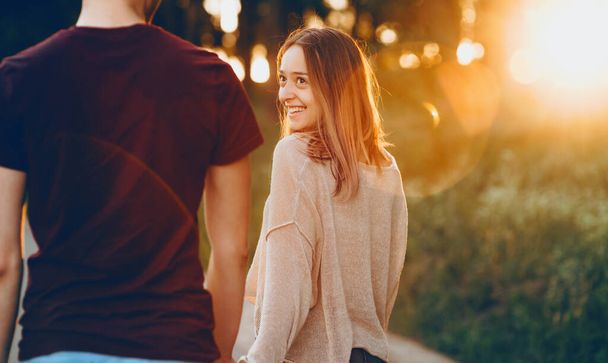Wesoła biała dziewczyna idzie ręka w rękę ze swoim kochankiem w parku podczas pięknego zachodu słońca - Zdjęcie, obraz