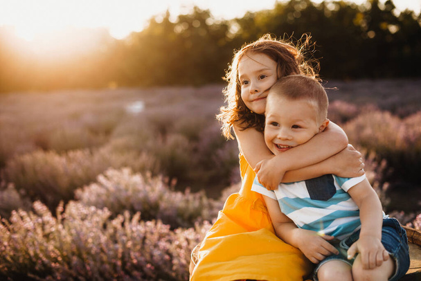 voorzichtige zus omarmen haar kleine broer terwijl poseren tegen de zon met een lavendel veld op de achtergrond - Foto, afbeelding