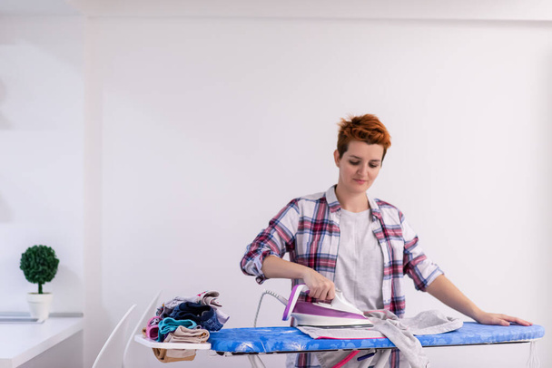 beautiful young red haired housewife in good mood ironing clothes on ironing board in front of a white wall at home - Foto, imagen
