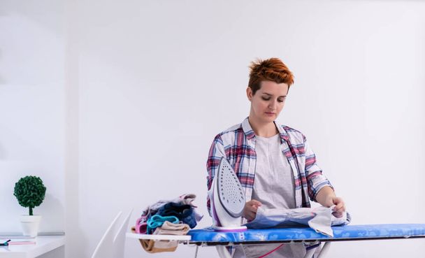 beautiful young red haired housewife in good mood ironing clothes on ironing board in front of a white wall at home - Foto, immagini