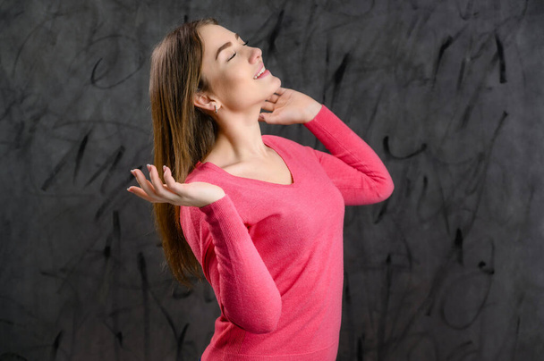 A woman model stands right in front of the camera. A pretty young Caucasian woman in a pink sweater on a gray background is talking and rejoicing. - Foto, afbeelding