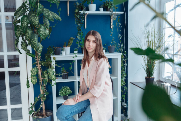 A girl in a pink sweater is sitting in the middle of the frame. In the foreground, the leaves of the plant are blurred. In the background are many houseplants. - Φωτογραφία, εικόνα