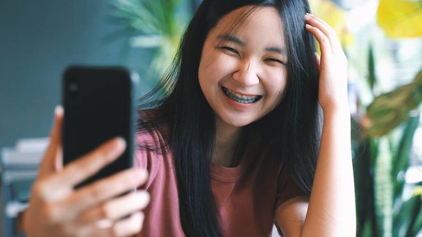 jong gelukkig aziatische duizendjarige vrouw het nemen van selfie en glimlachen met beugels in groen natuurlijk cafe, lifestyle concept - Foto, afbeelding