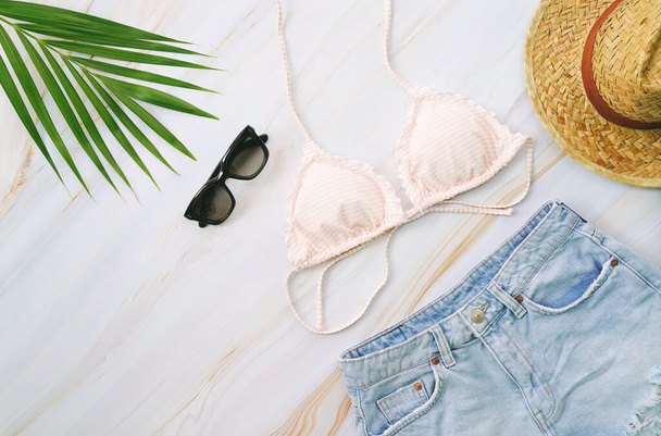 Płaskie ułożenie przedmiotów letnich z pastelowym bikini, okularami przeciwsłonecznymi, dżinsami, kapeluszem i zieloną rośliną tropikalną na marmurowym tle, modą i koncepcją lata - Zdjęcie, obraz