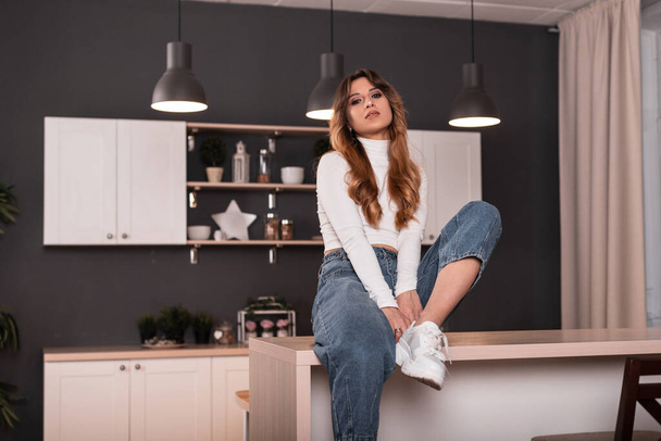 Cute cool young woman with long red hair in trendy youth casual blue-white wear posing sitting on a wooden table in the cook room. Sexy girl fashion model resting in the kitchen. Beauty stylish lady. - 写真・画像