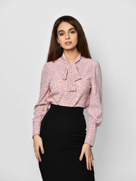 Young beautiful brunette woman is posing in formal pink blouse shirt and black skirt with her hands on hips - Fotografie, Obrázek