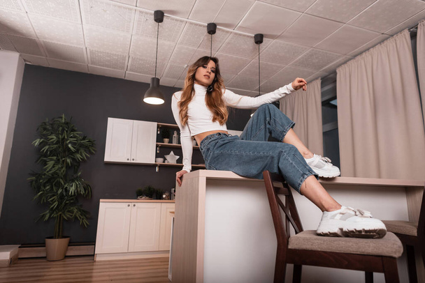 Das modische rothaarige Modell einer jungen Frau im schicken T-Shirt in blauen Vintage-Jeans in trendigen weißen Schuhen ruht auf einem Holztisch im Haus. Schön modern mädchen hausfrau im die küche zimmer. - Foto, Bild