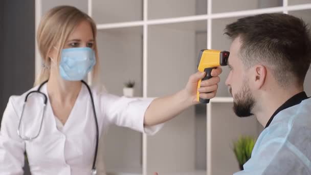 De arts in een witte jas en een medisch masker meet de temperatuur van een patiënt in het medisch kantoor met een infrarood contactloze thermometer. - Video