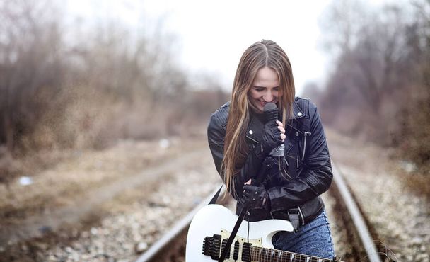 Όμορφη νεαρή κοπέλα κουνιστή πολυθρόνα με ηλεκτρική κιθάρα. Τραγουδά ένα ροκ μουσικός κορίτσι σε ένα δερμάτινο σακάκι με μια κιθάρα. Σολίστ ροκ μπάντα παίζει κιθάρα και κραυγάζει στο μικρόφωνο - Φωτογραφία, εικόνα