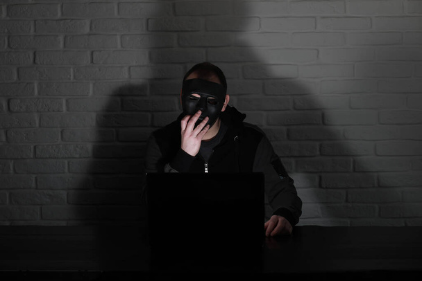 Siyah maskeli ve başlıklı bilgisayar korsanı Monito 'nun önündeki masada. - Fotoğraf, Görsel