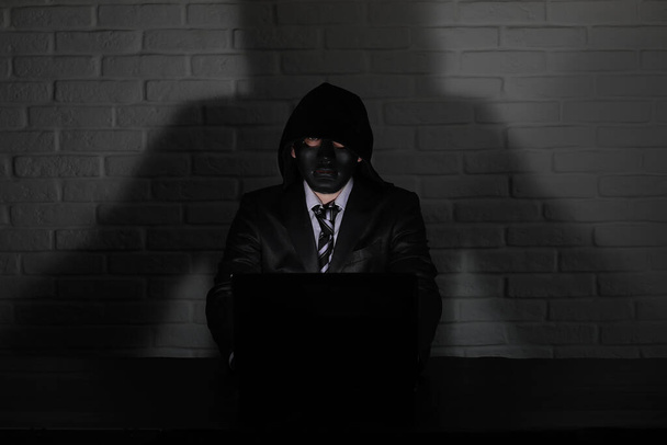 Hacker in black mask and hood at the table in front of the monito - Фото, изображение