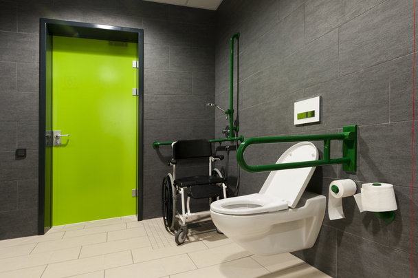 Um banheiro para pessoas com deficiência com barras verdes, cadeira de rodas e porta
 - Foto, Imagem